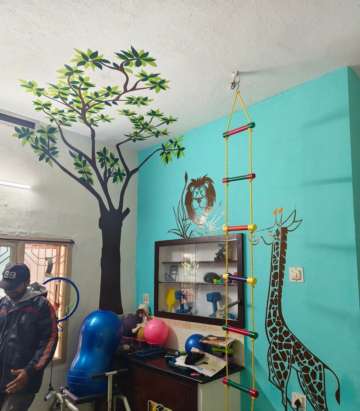 Play-based therapy room with tree and animal wall murals, a climbing rope ladder, gym balls and an equipment cabinet